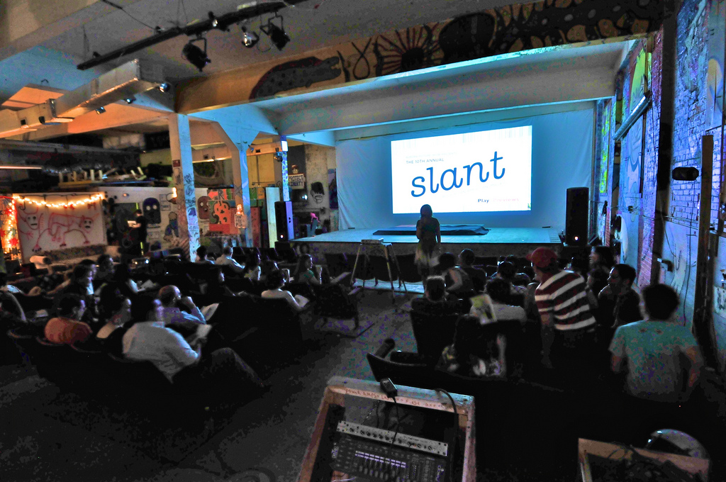 An audience inside an art space watching a film screening. The words "slant" are on the screen.