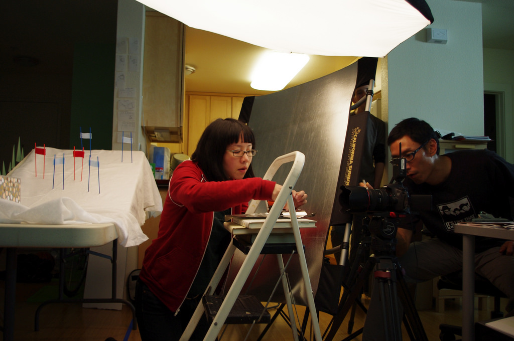 The set for a stop motion animation film. An Asian woman, the director, sets up the shot while an Asian man, the director of photography, adjusts the camera.