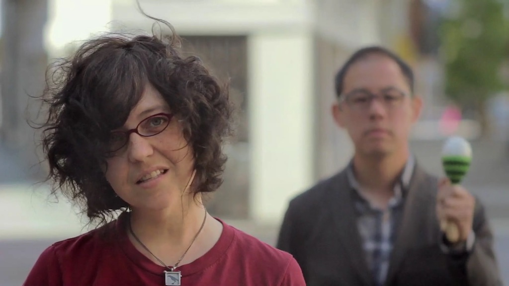 A still from a music video for The Invisible Cities. A white woman in a red shirt stands in the foreground. An Asian man stands in the background.