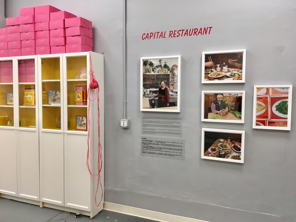 A photo exhibit inside a gallery. A few photos of Capital Restaurant are on the wall. There is also a cabinet with pink bakery boxes stacked on it.