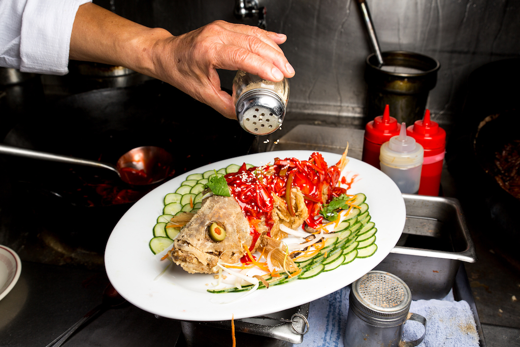 A cooked fish on a platter with cucumber slices artfully arranged around it. A hand holds a jar of seasoning over the platter. Photo by Michelle Min.