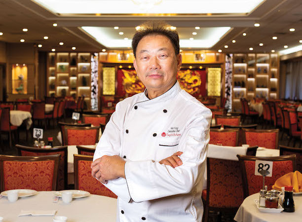 Chef Hoi Fung, wearing a white chef's jacket, in the dining room of his restaurant with round tables and red chairs.